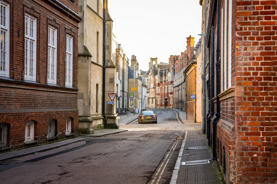 A narrow street in Kingston upon Thames lined with traditional brick and stone buildings, some with white-framed sash windows, and others with decorative brickwork. The street is paved with asphalt and features double yellow lines along the curb, indicating parking restrictions. A small, dark-coloured car is parked near the top of the slight incline, approaching a bend in the road. Bicycles are leaned against the buildings on the left side of the street, and a 'Give Way' road sign is visible on a pole near a building with a beige stone façade. The street is illuminated by soft natural light, suggesting late afternoon or early evening. The image captures the typical setting where house removals and furniture transport might encounter access issues on narrow, historic streets in Kingston upon Thames, highlighting the challenges of home relocation logistics in such environments, which Kingston upon Thames Removals can assist with.