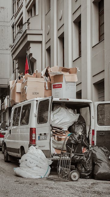Image of a white moving van parked on a narrow street in Kingston upon Thames, with the rear doors open revealing various packing materials, including large white plastic bags, cardboard boxes, and wrapped furniture. Several packed boxes are stacked on top of the van, some with labels, while others are without. A small trolley is positioned near the van, loaded with additional boxes and items, indicating an active home relocation process. The van is situated close to the façade of a multi-storey building with uniform windows and stone detailing, characteristic of urban residential architecture. The setting appears to be during daylight hours, with natural light illuminating the scene. This image reflects the logistical challenges of furniture transport and packing during a house removal, emphasizing the importance of careful loading and spatial management, as handled by Kingston upon Thames Removals for their removals services.