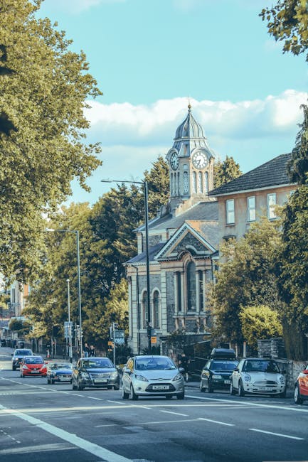 A narrow cobbled street lined with traditional red brick buildings and architectural details such as bay windows, decorative brickwork, and iron railings. A white van is parked on the right side, like