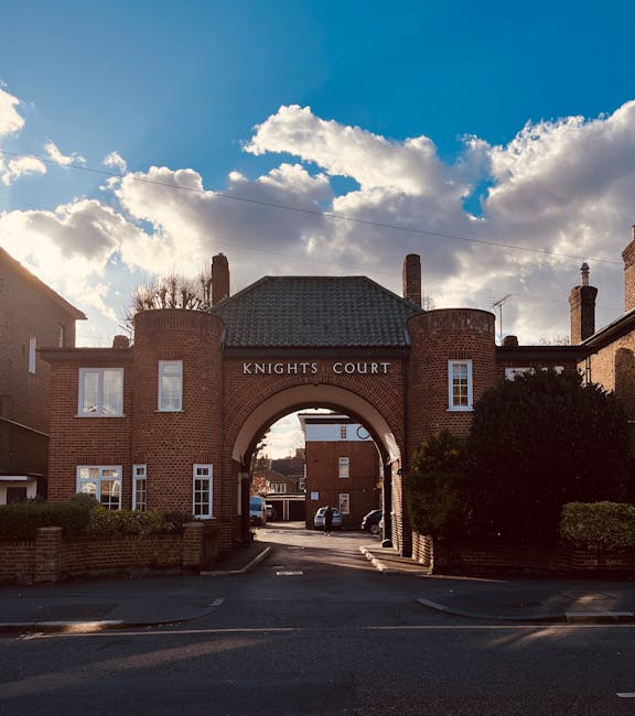 Photograph of the entrance to Knights Court, a residential building with a prominent brick archway leading to a courtyard or parking area. The building features brick walls, white-framed windows, and a tiled gable roof. Above the arch, there is a rounded section of the building with the inscription 'KNIGHTS COURT'. The sky above is partly cloudy with patches of blue, and the sunlight casting shadows on the brickwork. To the left and right of the entrance, there are bushes and trees, adding greenery to the scene. The image captures the exterior of the property, illustrating a typical residential setting relevant to house removals and moving services, with a focus on urban living spaces often involved in home relocation and furniture transport in Kingston upon Thames.