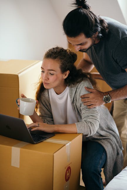 Interior scene showing a woman seated at a table surrounded by several large cardboard moving boxes, some opened and sealed with packing tape, with a laptop in front of her. She holds a white ceramic mug in one hand and appears focused on the laptop screen. A man stands close beside her, leaning over and placing a hand gently on her shoulder while looking at the laptop. The room features neutral-colored walls and natural lighting, suggesting a home environment preparing for or in the process of a house move. The setting highlights packing and home relocation activities, emphasizing the importance of organized furniture transport and careful handling during the moving process as managed by Kingston upon Thames Removals.