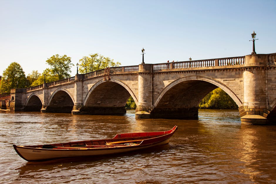 A red and white rowing boat floating on a river in front of a stone bridge with multiple arches, decorative lampposts along the bridge's railing, and lush green trees in the background. The scene is b