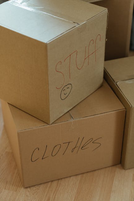 Close-up image of several brown cardboard boxes stacked together on a wooden floor, used for home relocation. The top box has the handwritten label 'STUFF' in red marker with a simple smiling face dra