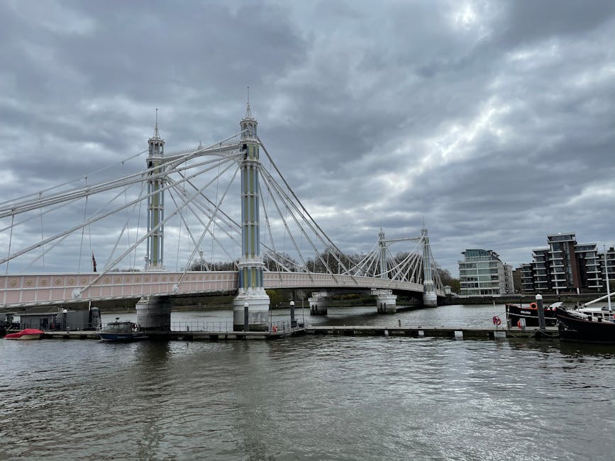 A view of a large suspension bridge spanning a river, with two tall towers supporting steel cables and a pink and white painted deck. The bridge is situated in an urban environment with modern residential buildings visible on the right bank of the river. The water below is calm, with small boats docked at the shoreline. Overhead, the sky is filled with dark, heavy clouds, suggesting an impending storm. This scene captures an iconic London bridge close to Kingston Bridge in Kingston upon Thames, with a backdrop suitable for home relocation and furniture transport activities facilitated by Kingston upon Thames Removals, evidenced by the surrounding setting and structural features.