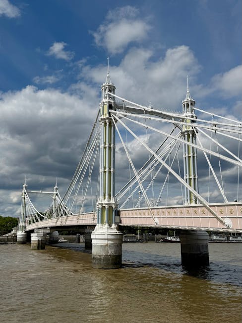 A wide-angle view of the Hammersmith Bridge spanning the River Thames on a partly cloudy day, with large stone pillars supporting the bridge structure. The bridge features a pale pink painted railing with decorative elements, and the suspension cables are anchored to tall, ornate metal towers with pointed finials. The water beneath appears calm with gentle ripples, and in the background, the sky is filled with a mix of dark and light clouds, creating a dramatic atmosphere. This scene depicts a historic suspension bridge located in London, and could relate to transport or infrastructure themes relevant to moving services or city logistics. Kingston upon Thames Removals, a professional moving company, may occasionally work with locations near iconic bridges such as this one during home relocation or furniture transport projects, utilizing careful handling and loading procedures near bridges and urban infrastructure.
