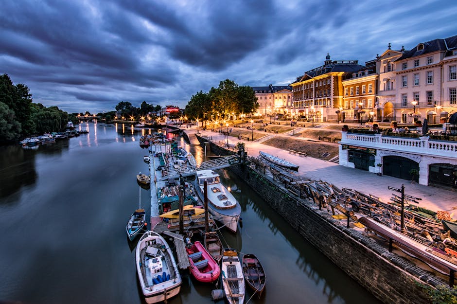 Aerial view of a riverside area at dusk, showing multiple boats docked along a calm waterway with reflections of the boats and illuminated buildings lining the street. The scene features a row of large, elegant white and pastel-coloured buildings with decorative lighting and outdoor seating areas, situated on a paved promenade. The sky overhead is dark with heavy clouds, contrasting with the warm glow of streetlights and building lights. The surroundings include lush green trees and a distant bridge visible over the river. This scene illustrates the typical environment for a home relocation or furniture transport process near Kingston Riverside, with potential loading areas on the pavement for moving vans as used by professionals like Kingston upon Thames Removals during packing and furniture transport activities.
