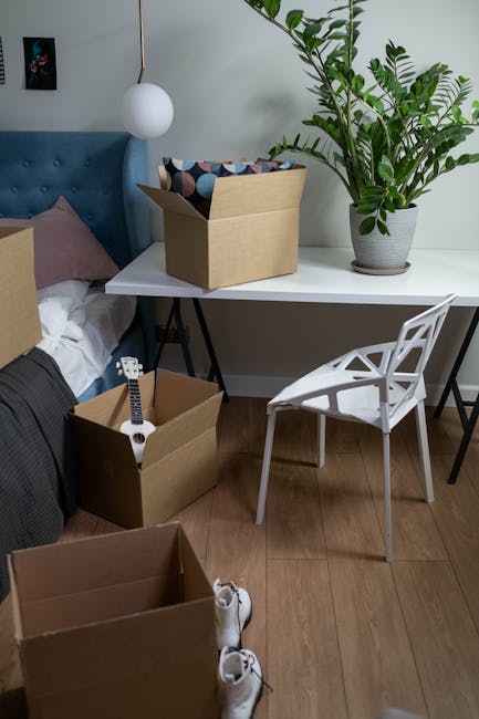 A room scene showing several packing boxes, including a cardboard box on a white desk containing colorful cushions and a large potted plant with broad green leaves positioned on the desk. Another open box on the floor contains a small white toy guitar and additional packing materials, while a pair of white sneakers is placed nearby on the wooden floor. A white modern chair with geometric cutouts is situated next to the desk. The bed, partially visible on the left, is covered with white and dark bedding, and a soft pink cushion is resting against a teal upholstered headboard. There is a wall-mounted spherical white pendant light above the bed, and a small framed picture hanging on the light-colored wall. The scene depicts a home during a packing process, possibly related to home relocation or furniture transport, as managed by Kingston upon Thames Removals.