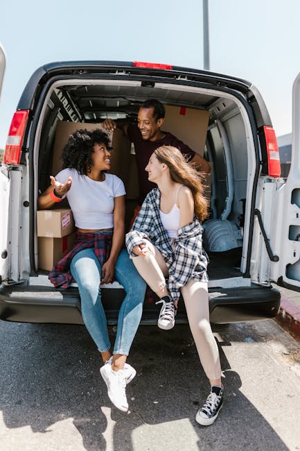 A group of three young adults sitting inside the open rear of a white moving van parked outdoors on a bright day. The van's interior shows packed cardboard boxes and some wrapped furniture or household items. The individuals are engaged in conversation, smiling and gesturing, with some boxes visible in the background. The scene captures the loading or unloading process during a home relocation, supported by the truck exhibiting a spacious cargo area filled with packing materials, ready for furniture transport or moving services. The environment appears to be on a residential street, with the pavement, a curb, and a clear sky overseeing the activity. Kingston upon Thames Removals specializes in professional removals, including packing, loading, and transporting household belongings, as reflected by the scene of furniture and boxes being managed during a house move.