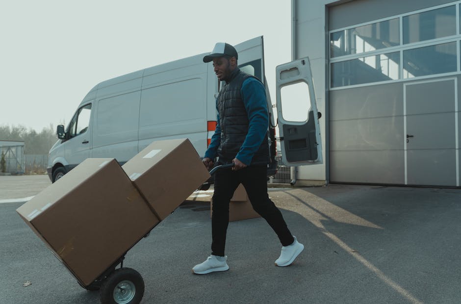 A man wearing a dark blue jacket, black pants, white sneakers, and a grey cap is seen loading two large cardboard boxes onto a hand truck outside a modern building with large glass windows and a grey sliding door. The hand truck is positioned on an asphalt surface, and the man is pushing it toward a white commercial van with its side door open, revealing the vehicle's interior ready for furniture transport or packing during a home relocation. Several additional cardboard boxes are stacked nearby on the ground. The scene is set in daylight, with natural light illuminating the area, and the background includes a partly cloudy sky and some distant trees, indicating an organized moving process managed by Kingston upon Thames Removals for a residential or commercial move in the KT6 area.