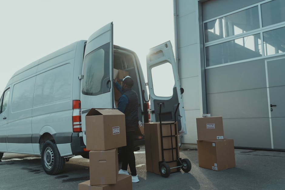 A group of three young adults sitting inside the open rear of a white moving van parked outdoors on a bright day. The van's interior shows packed cardboard boxes and some wrapped furniture or household items. The individuals are engaged in conversation, smiling and gesturing, with some boxes visible in the background. The scene captures the loading or unloading process during a home relocation, supported by the truck exhibiting a spacious cargo area filled with packing materials, ready for furniture transport or moving services. The environment appears to be on a residential street, with the pavement, a curb, and a clear sky overseeing the activity. Kingston upon Thames Removals specializes in professional removals, including packing, loading, and transporting household belongings, as reflected by the scene of furniture and boxes being managed during a house move.